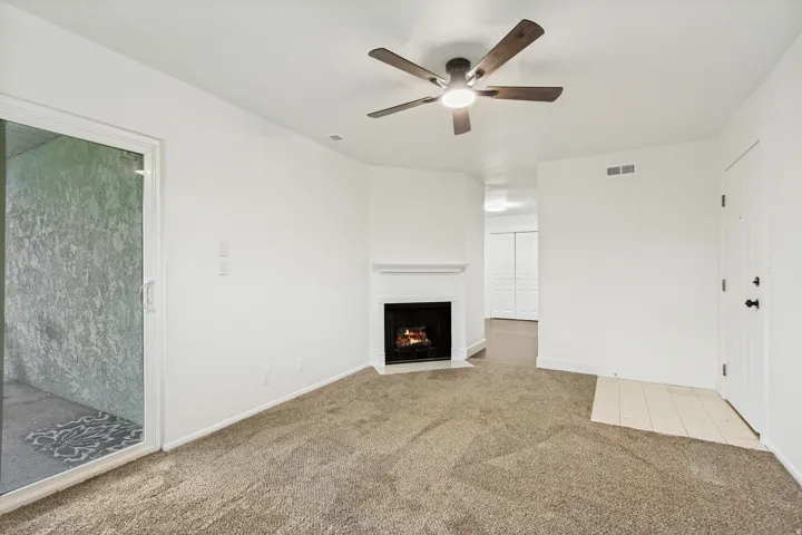 Living area featuring a corner fireplace with white mantel, sliding glass door, overhead ceiling fan with light, and neutral carpet flooring
