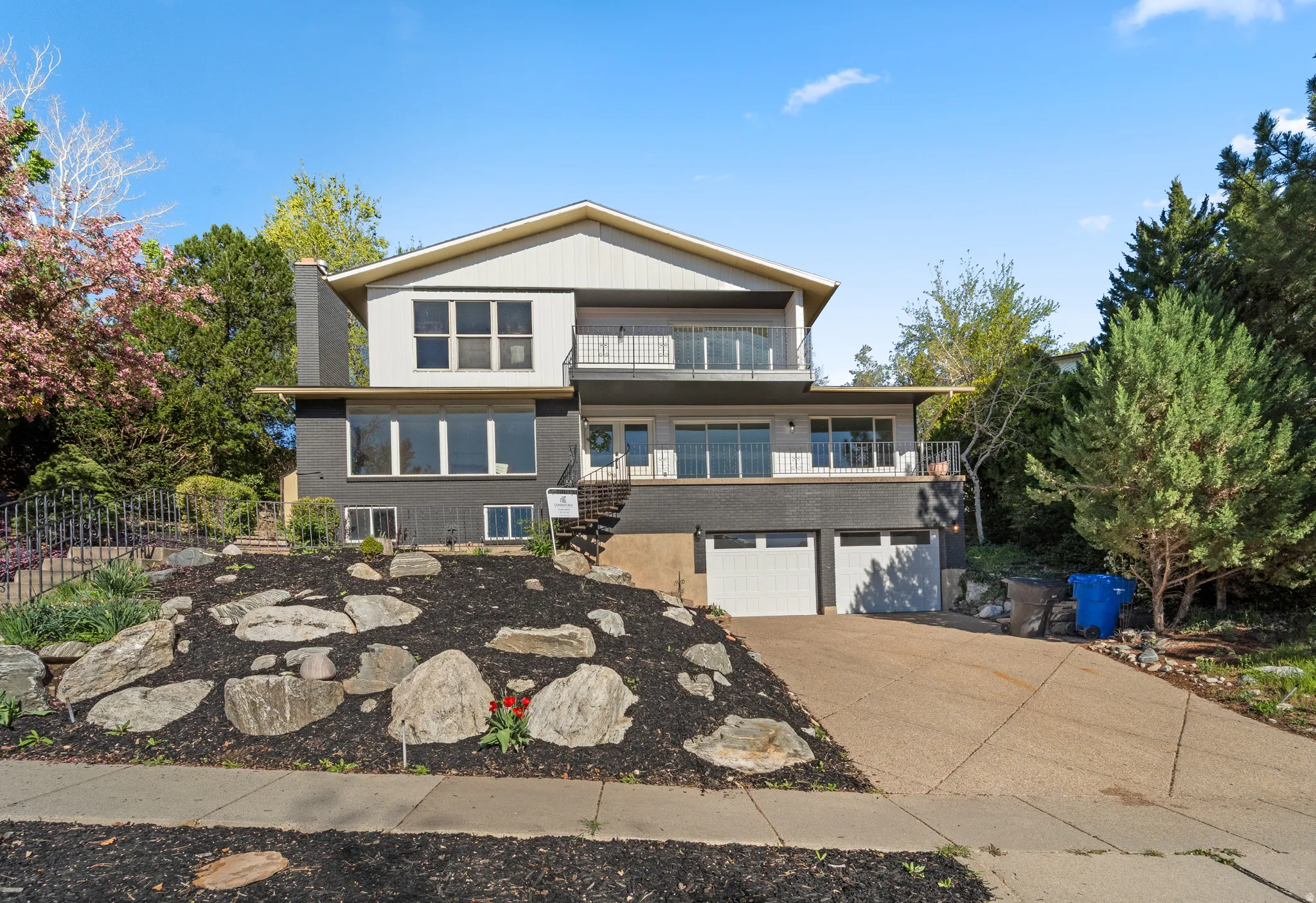 Multi-level residence featuring a two-car garage, multiple balconies with wrought iron railings, a prominent chimney, and a rock-landscaped front yard with mature trees