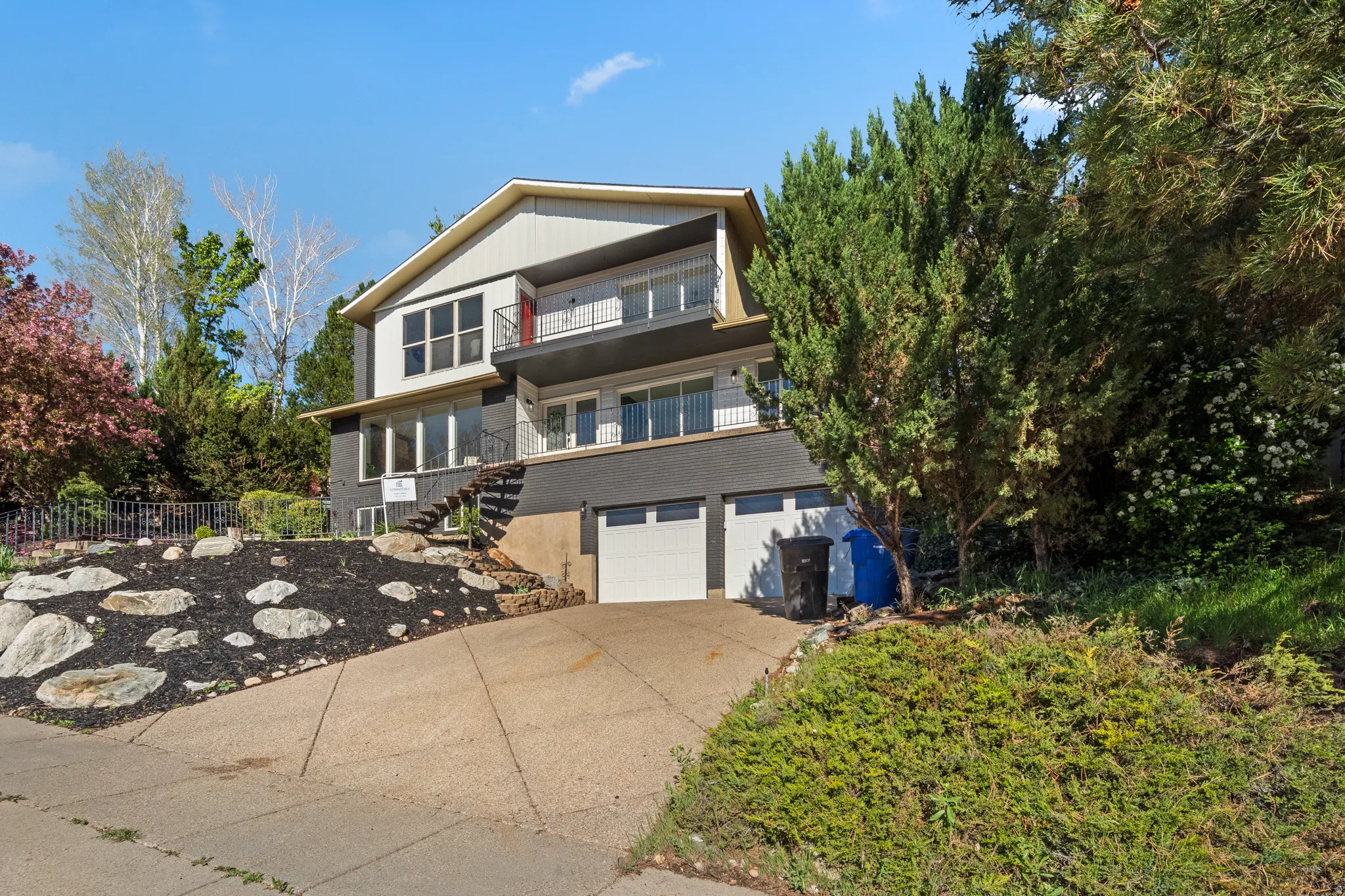 Multi-level residence featuring a two-car garage with white doors, a tiered front landscaping with rocks and dark ground cover, and mature trees