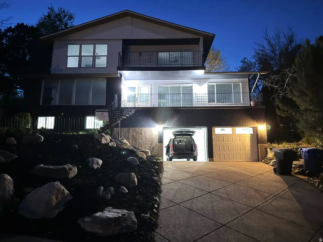 Multi-level exterior with a two-car garage, upper and lower balconies with black railings, and a landscaped rock garden