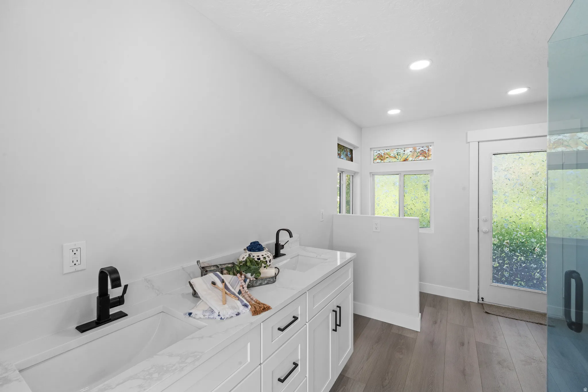 Dual vanity with integrated sinks and black matte faucets, white shaker cabinetry with black pulls, light wood-finish flooring, recessed lighting, and a frosted glass door