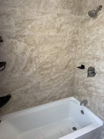Bathtub and shower combination featuring neutral-toned tiled walls with veining patterns