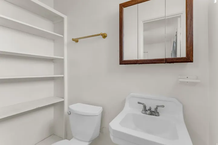 Compact bathroom featuring a wall-mounted sink, a toilet, built-in shelving, a wood-framed medicine cabinet, and a gold-finish towel bar