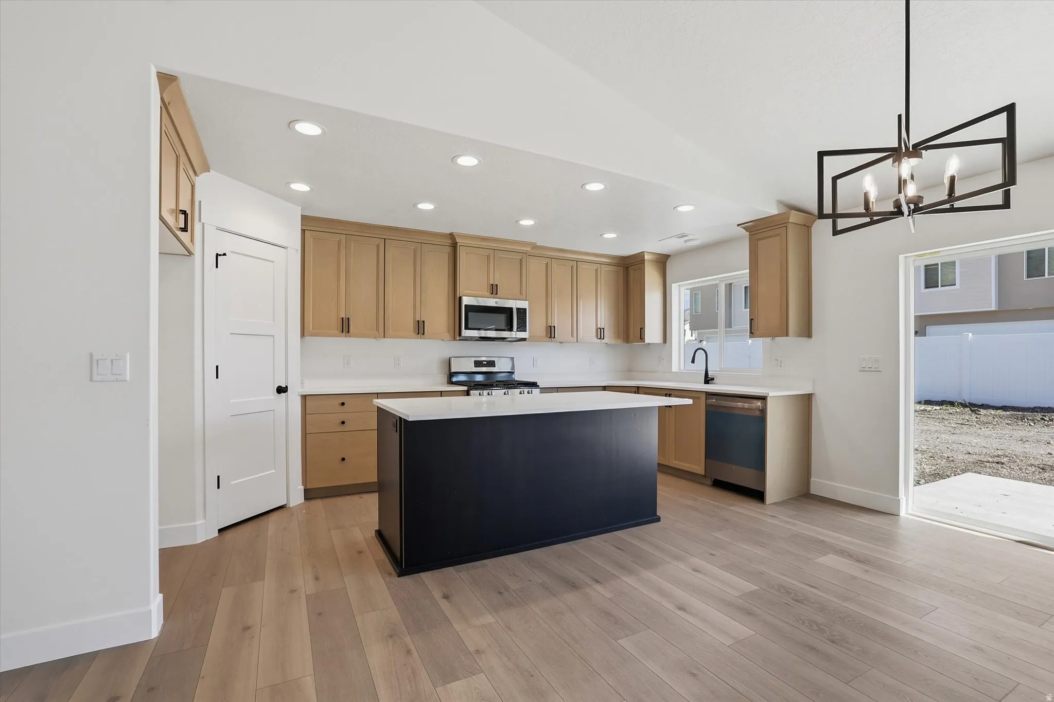 Modern kitchen featuring a dark-toned island with white countertop, light wood-finish cabinetry, stainless steel appliances, and wood-finish flooring