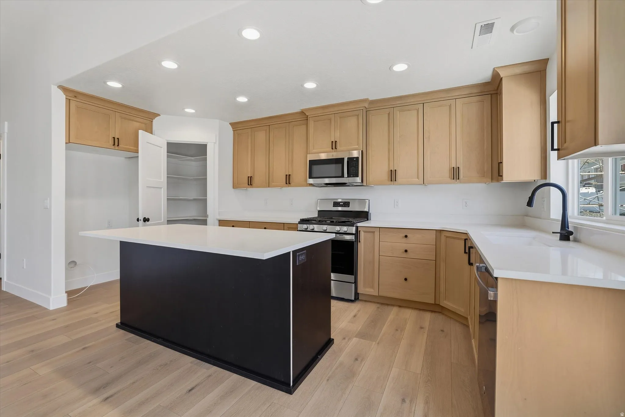 Kitchen featuring a central island with a light-toned countertop, wood-finish flooring, light wood cabinetry, recessed lighting, and stainless steel appliances