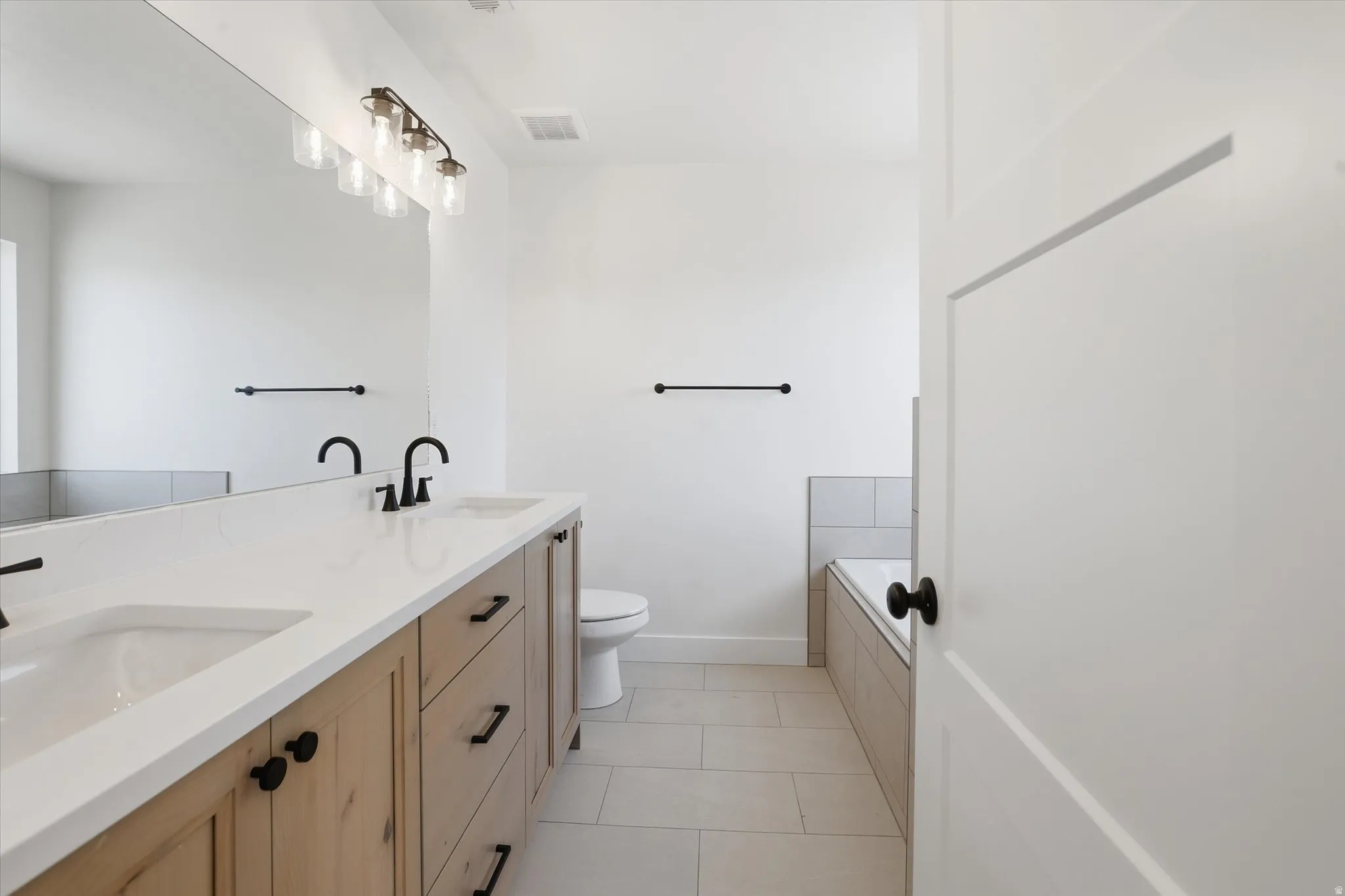 Bathroom featuring a dual-sink vanity with light wood-finish cabinetry, white countertops, and matte black fixtures