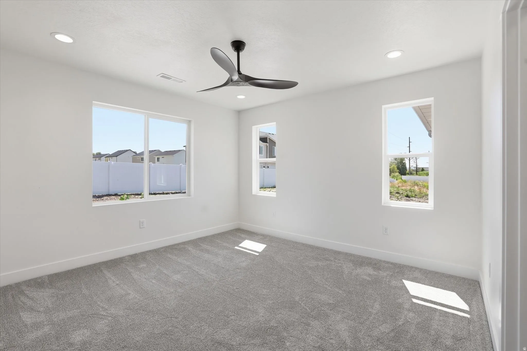 Spacious room featuring light gray carpeting, crisp white walls, multiple windows providing natural illumination, recessed lighting, and a modern ceiling fan