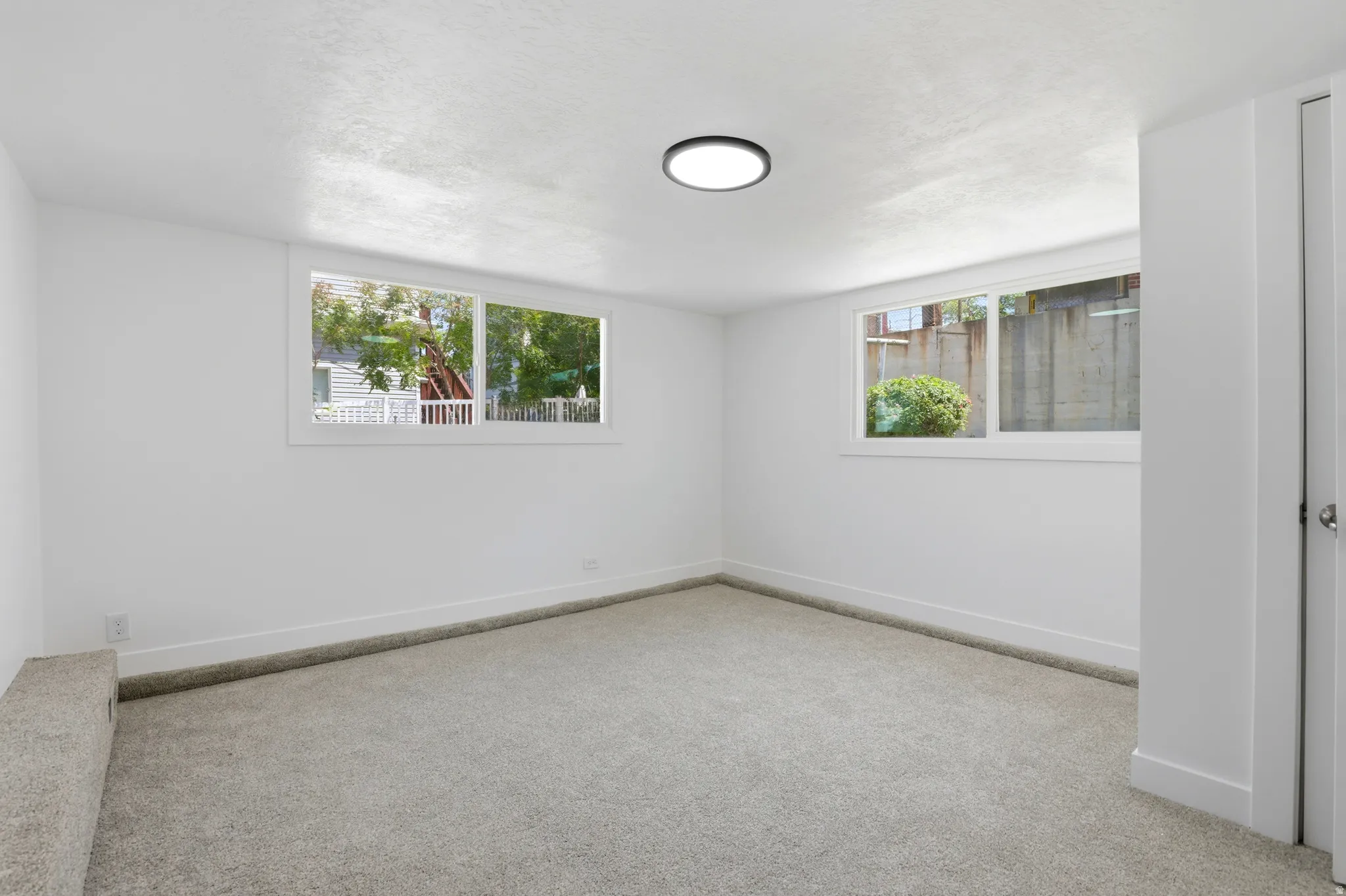 Spacious room featuring light gray carpeting, crisp white walls, multiple windows, a modern flush-mount ceiling light, and white baseboards