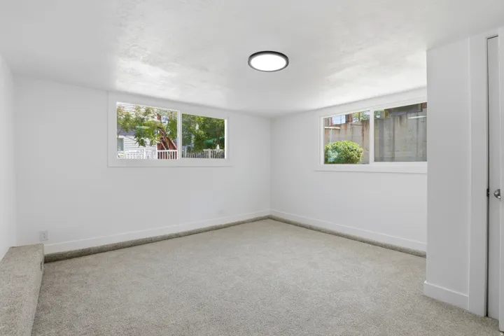 Spacious room featuring light gray carpeting, crisp white walls, multiple windows, a modern flush-mount ceiling light, and white baseboards
