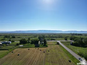 Expansive agricultural field with cut hay bales, surrounded by green foliage, a paved road, and distant mountain ranges under a clear sky