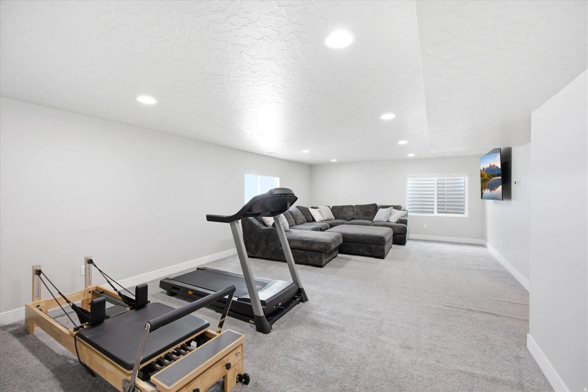 Spacious recreational room featuring light gray carpeting, recessed ceiling lighting, a wall-mounted display, and a window with white louvered blinds