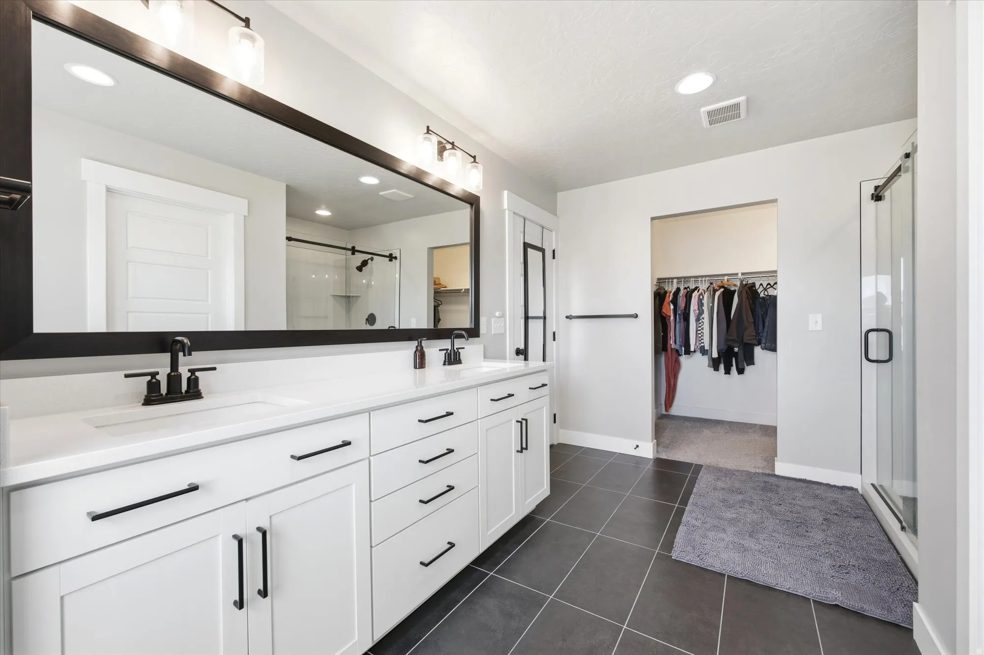 Spacious bathroom featuring a dual vanity with white cabinetry and black hardware, a large framed mirror, dark tile flooring, and a glass-enclosed shower