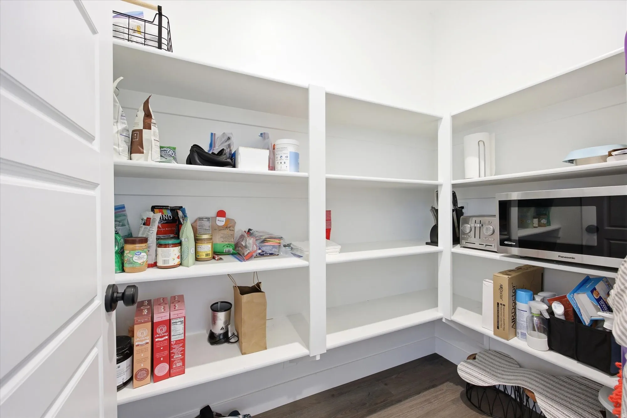 Spacious pantry featuring extensive white built-in shelving, dark wood-finish flooring, and a stainless steel microwave