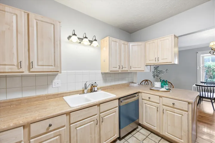 Kitchen featuring light wood-finish cabinetry, white tile backsplash, a double basin sink, and an integrated dishwasher