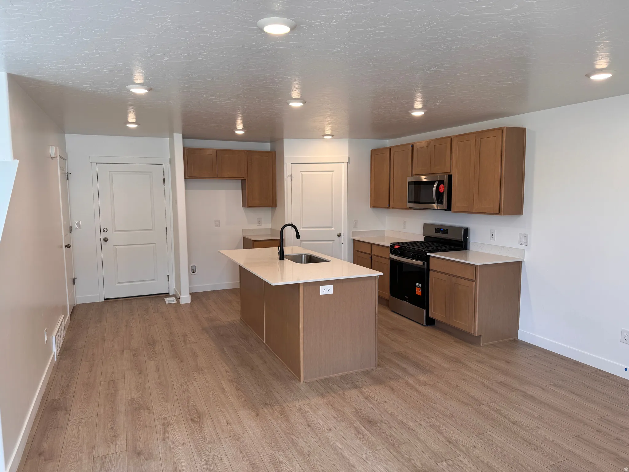 Modern kitchen featuring light wood-finish cabinetry, stainless steel appliances, a central island with an integrated sink, white countertops, and recessed lighting