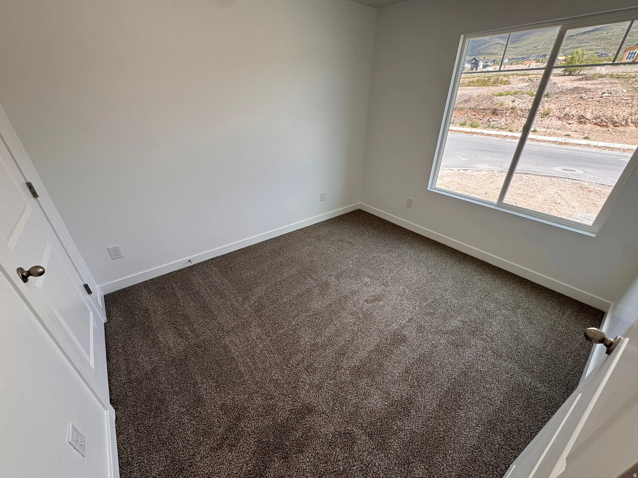 Interior room featuring white walls, dark carpet, a multi-pane window, and a white interior door with bronze hardware