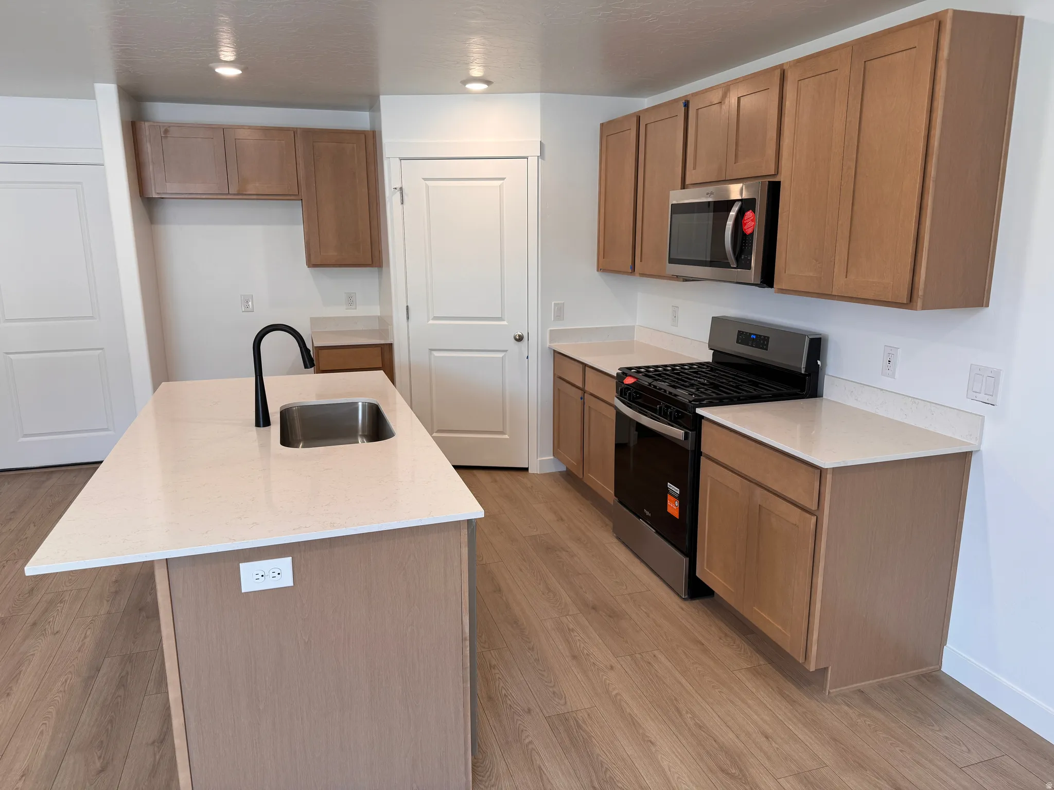 Kitchen featuring light wood-finish cabinetry, light-toned countertops, a central island with an integrated sink and black faucet, and wood-finish flooring