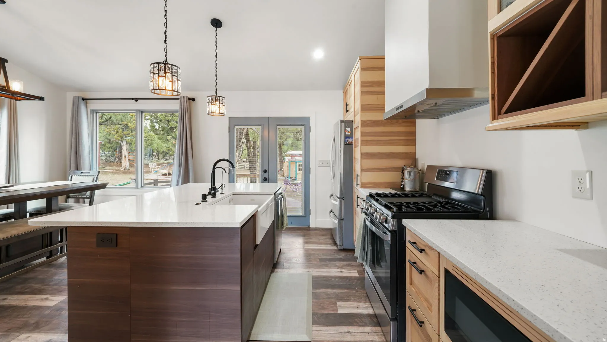Open-concept kitchen featuring a spacious island with white countertop and farmhouse sink, stainless steel appliances, wood-finish flooring, and natural wood cabinetry