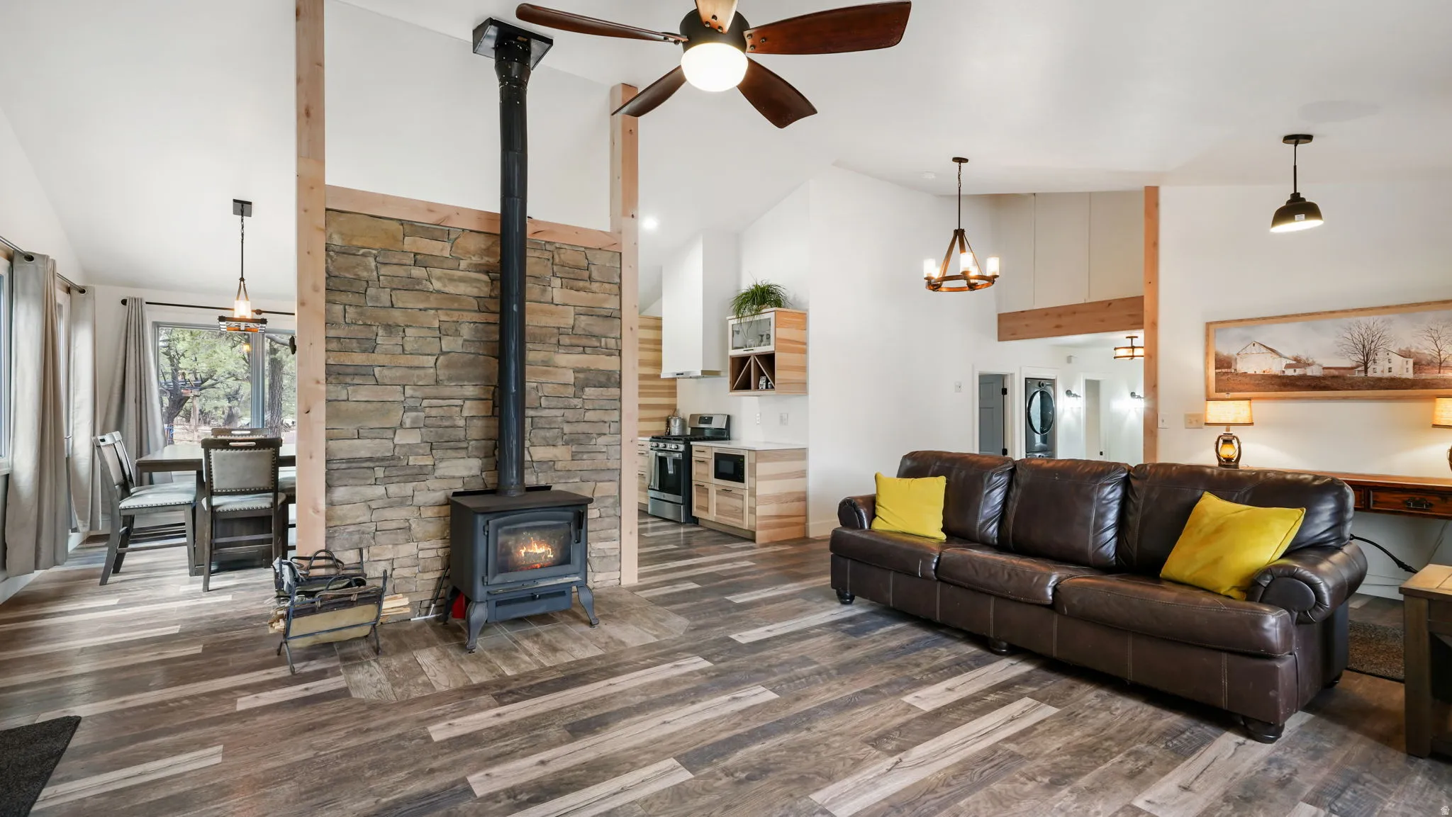 Expansive living area featuring wood-finish flooring, a stone-accented wall with a black wood-burning stove, vaulted ceilings, and a ceiling fan