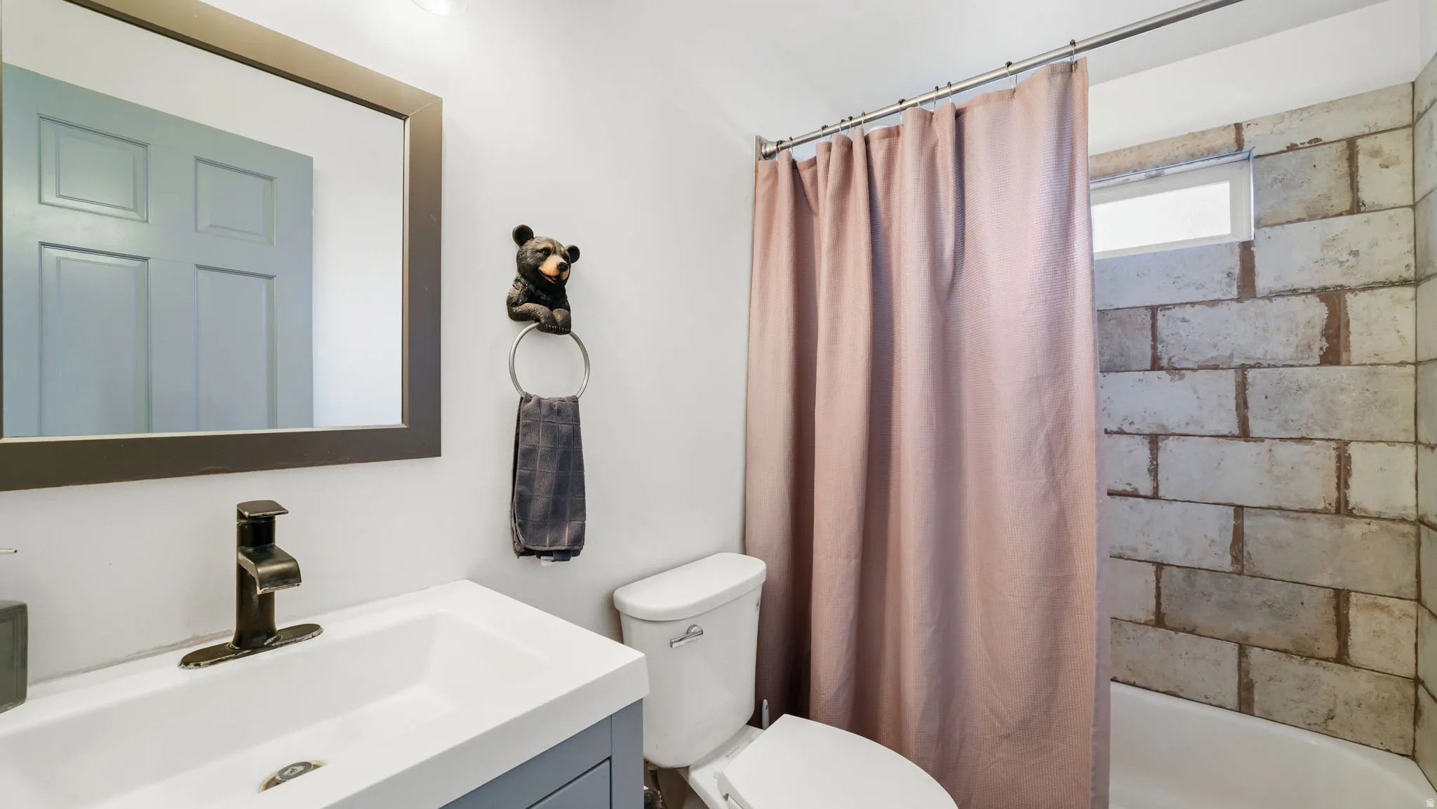 Bathroom featuring a single vanity with a white basin and dark-toned faucet, framed mirror, toilet, and a shower/tub combination with a light-toned brick-patterned tile surround