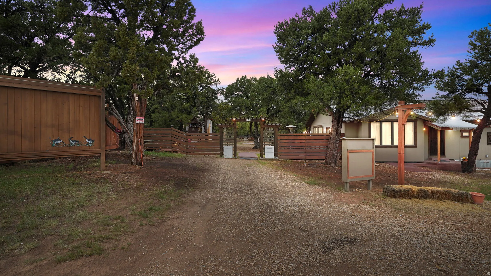 Gravel driveway entry with a wooden privacy fence, mature trees, and a visible portion of a single-story residence