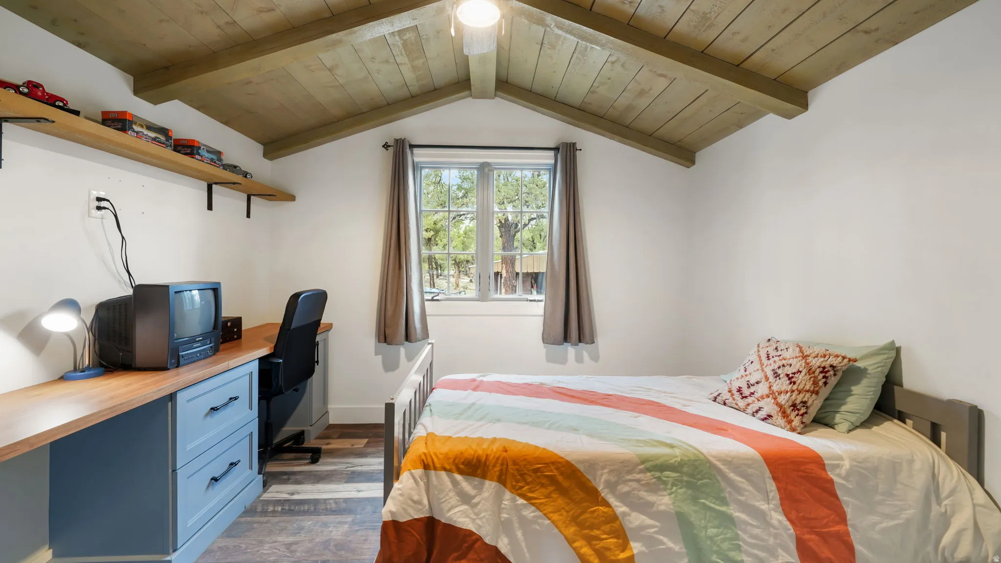 Vaulted wood-plank ceiling with exposed beams, white painted walls, wood-finish flooring, built-in desk with blue cabinetry, and a window with natural light
