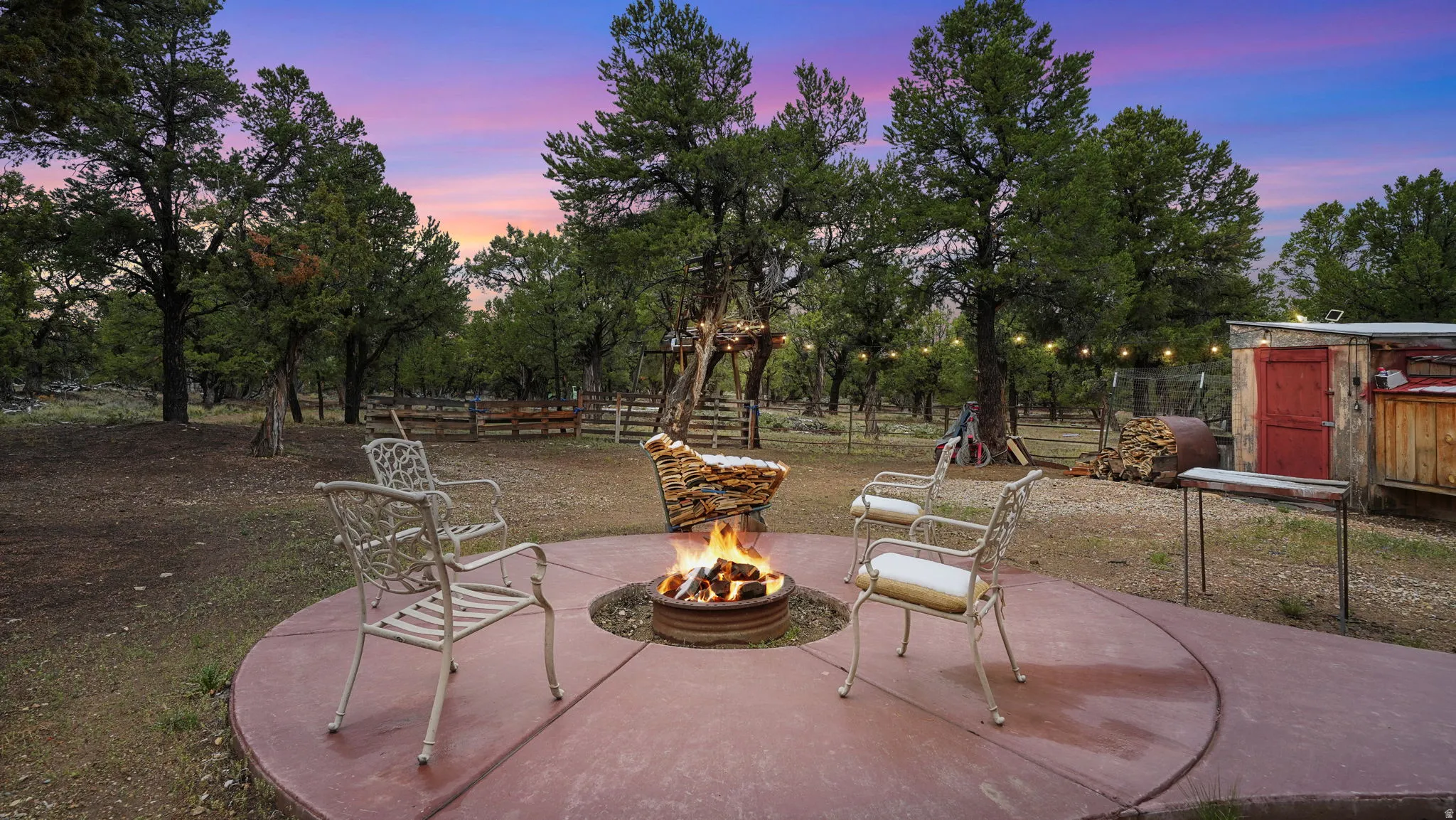 Outdoor concrete patio featuring a built-in fire pit, surrounded by mature trees, string lighting, a rustic shed, and a wooden fence line