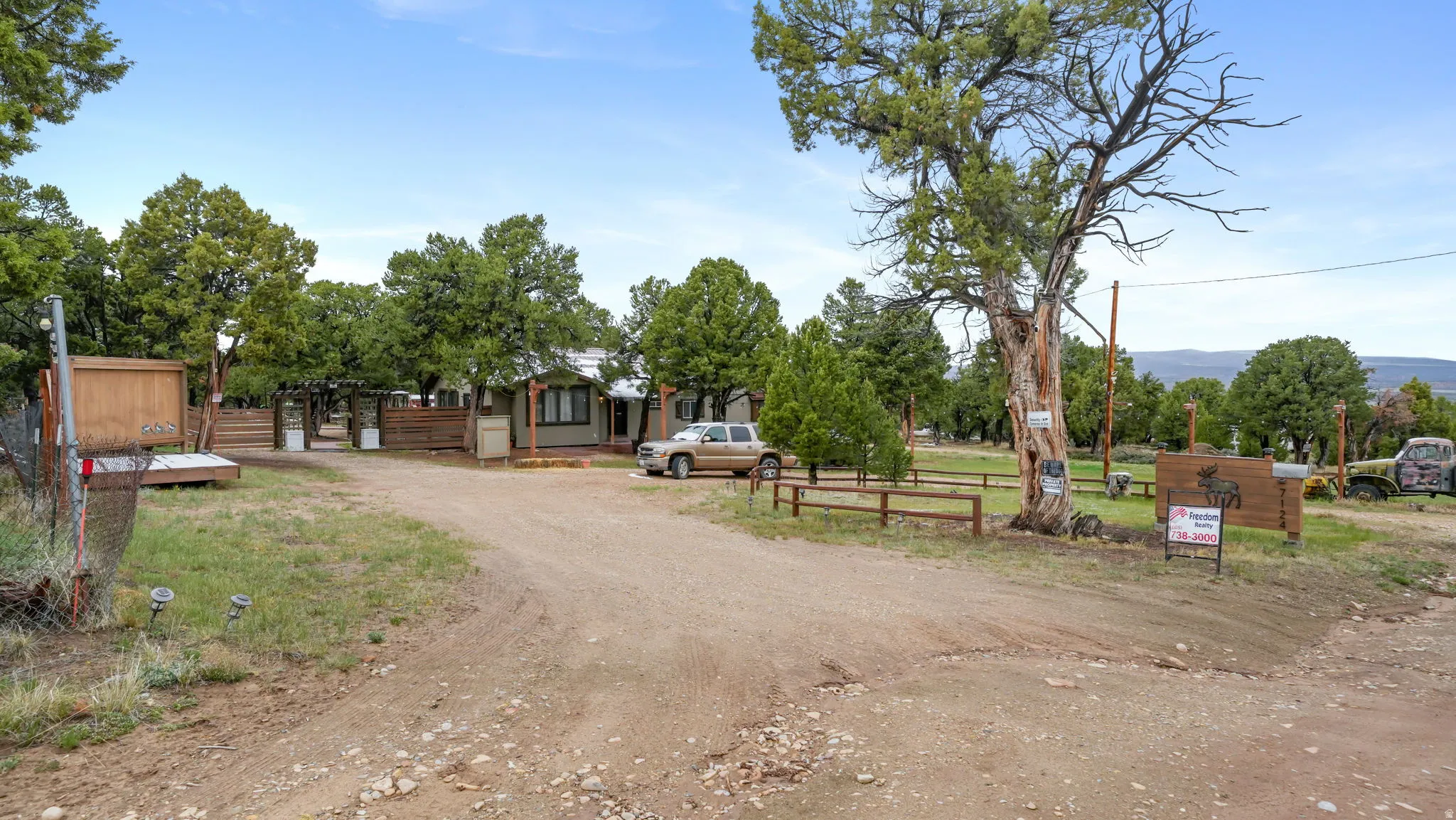 Gravel driveway leading to a residence featuring a covered front porch, mature trees, wood fencing, and a natural landscape