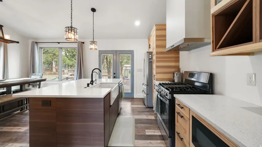 Open-concept kitchen featuring a spacious island with white countertop and farmhouse sink, stainless steel appliances, wood-finish flooring, and natural wood cabinetry