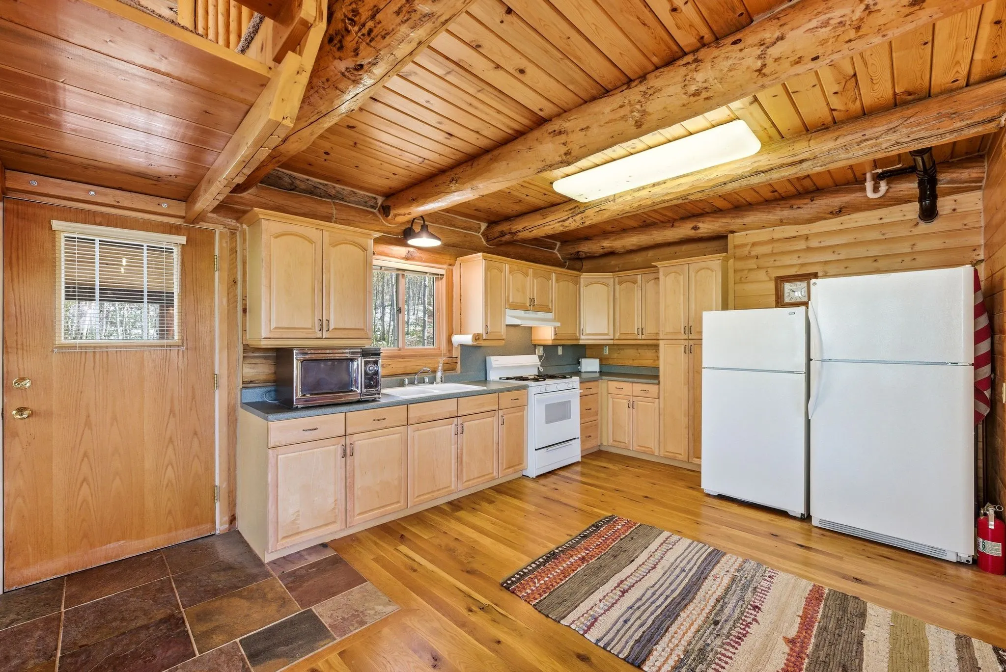 Rustic kitchen featuring natural wood walls and ceiling beams, light wood cabinetry, white appliances, and mixed flooring with wood-finish and stone tiles