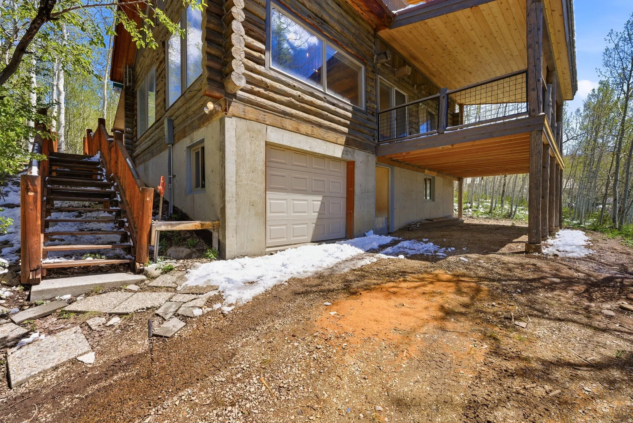 Rustic log home exterior featuring a concrete foundation, a ground-level garage with a panel door, a wood deck with a metal grid railing, and a log staircase with stone risers
