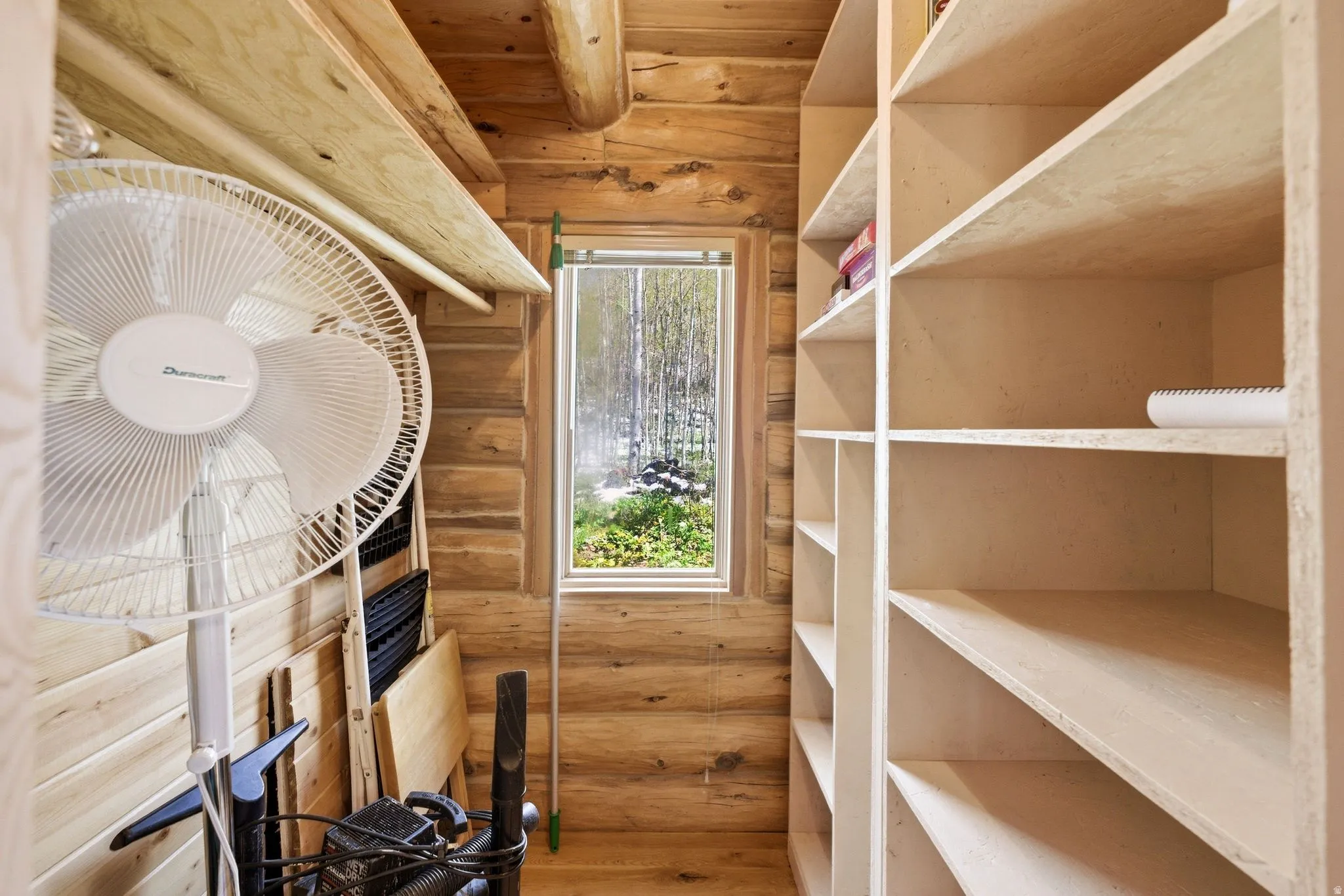 Storage area featuring extensive built-in shelving, natural wood walls, wood-finish flooring, a wood-framed window, and exposed wood ceiling beams
