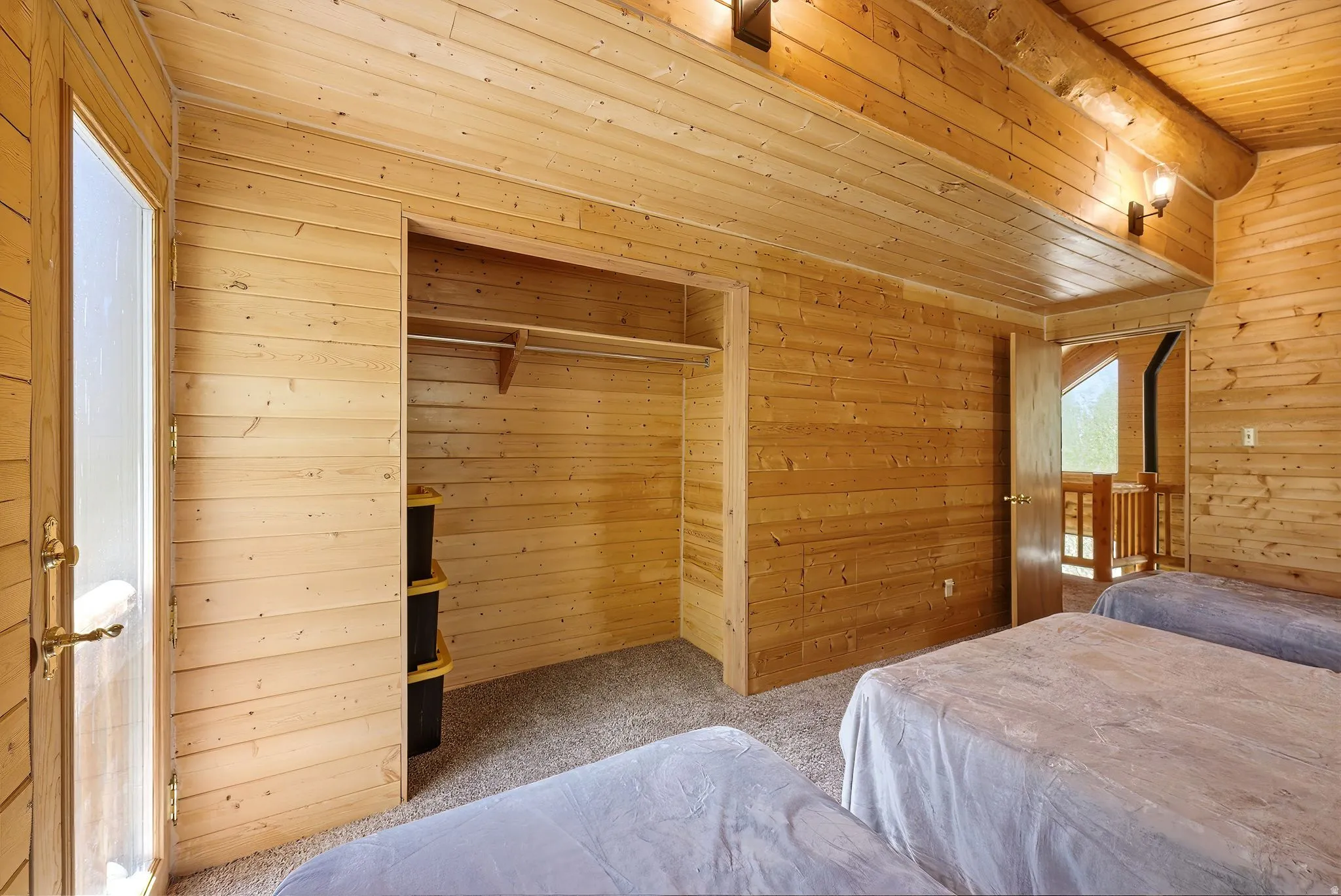 Room featuring wood plank walls and ceiling, carpet flooring, open closet with hanging rod and shelving, brass door hardware, and wall-mounted light fixtures