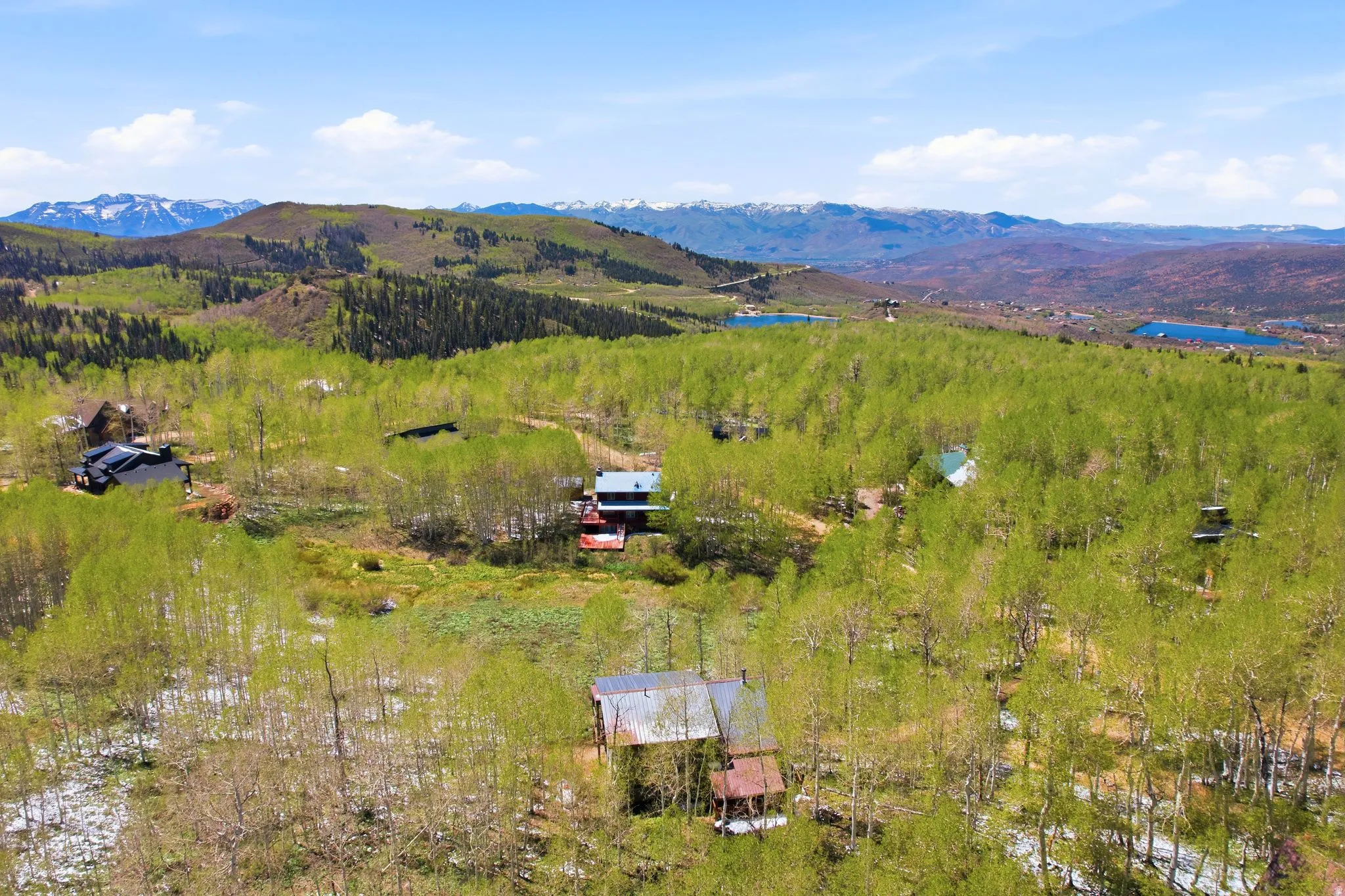 Mountainous landscape featuring several residences nestled among abundant green foliage, with distant snow-capped peaks and visible bodies of water