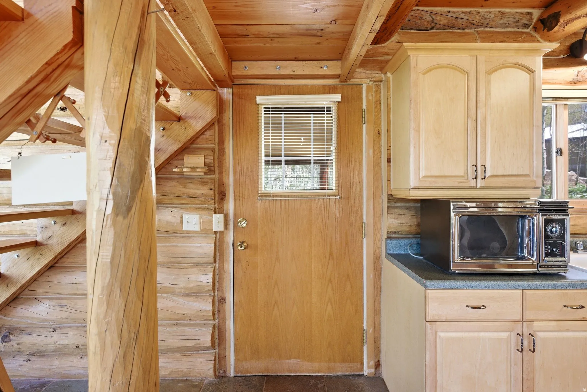 Wood-paneled interior featuring a solid wood door with window blind, a wooden staircase, and exposed log structural elements