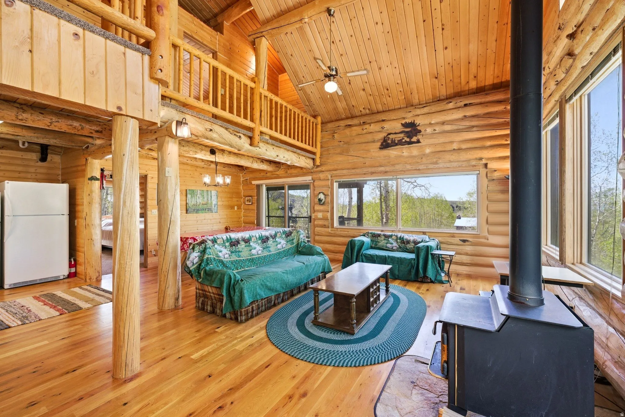 Expansive great room featuring log construction, wood-finish flooring, a wood-burning stove on a stone hearth, and a vaulted wood-paneled ceiling with a fan