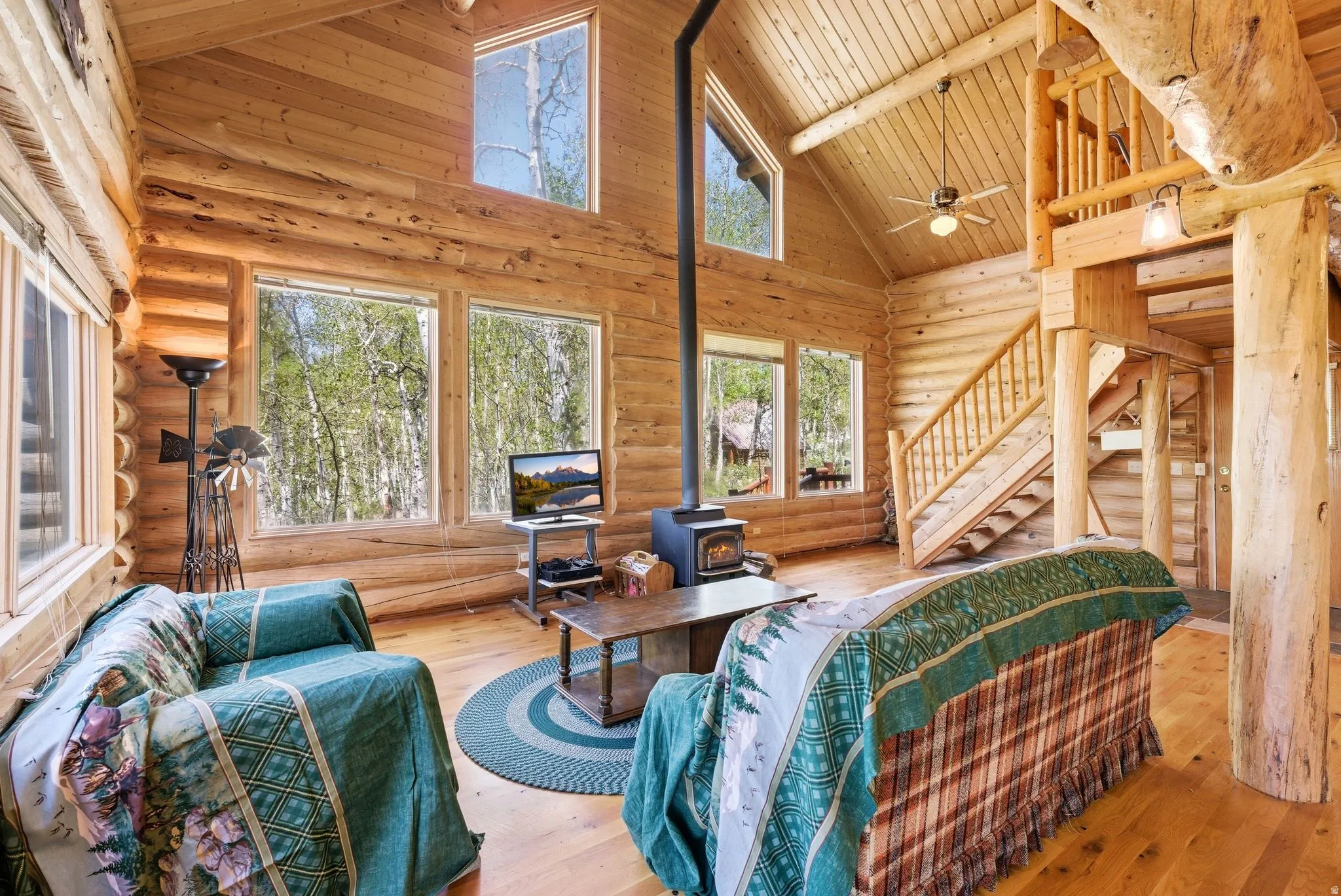Vaulted ceiling living area featuring log wall construction, natural wood-finish flooring, a wood-burning stove with a metal flue, and a prominent wood staircase leading to an upper level