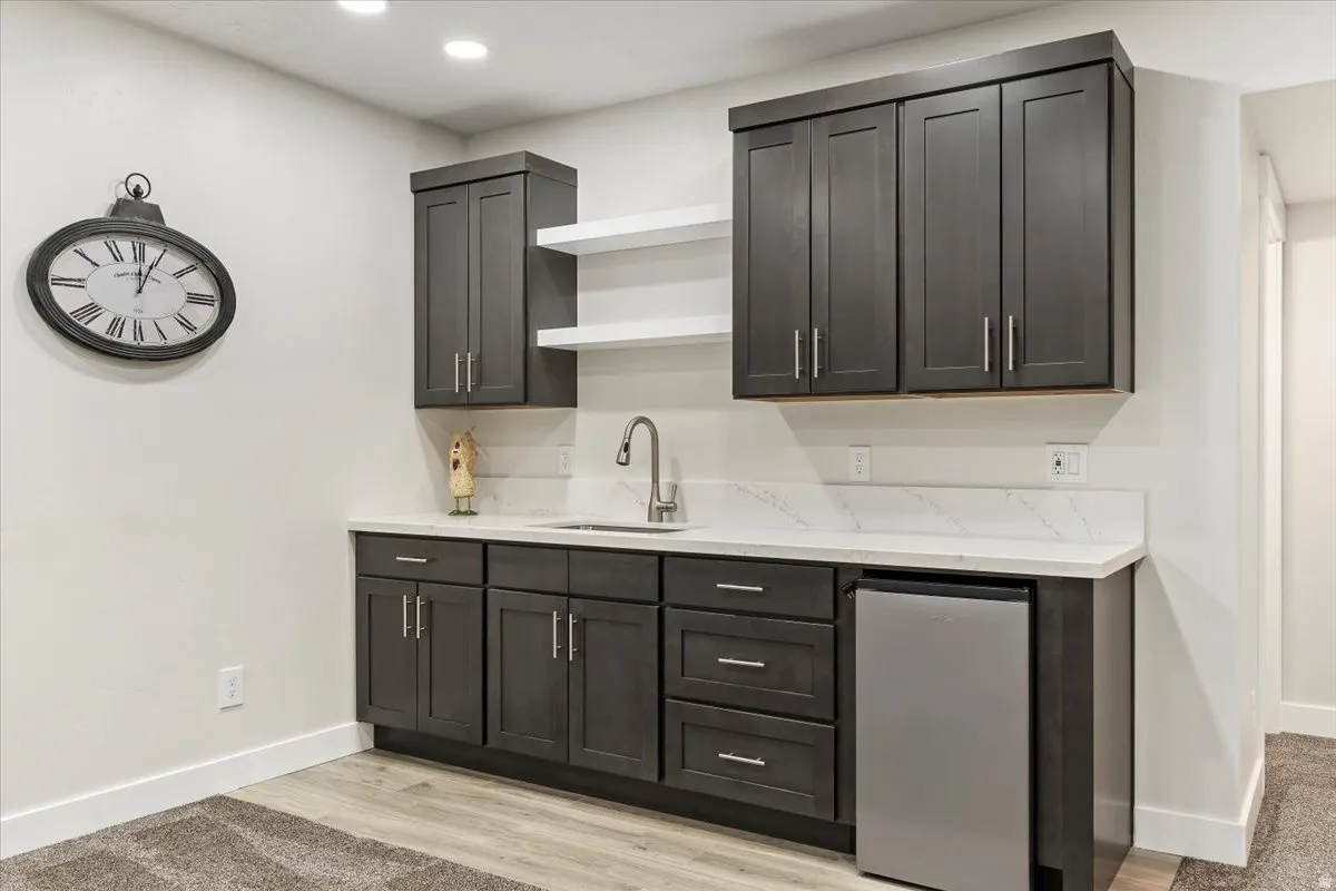 Built-in wet bar featuring dark cabinetry, a stainless steel sink, a modern faucet, quartz countertops, and a compact refrigerator