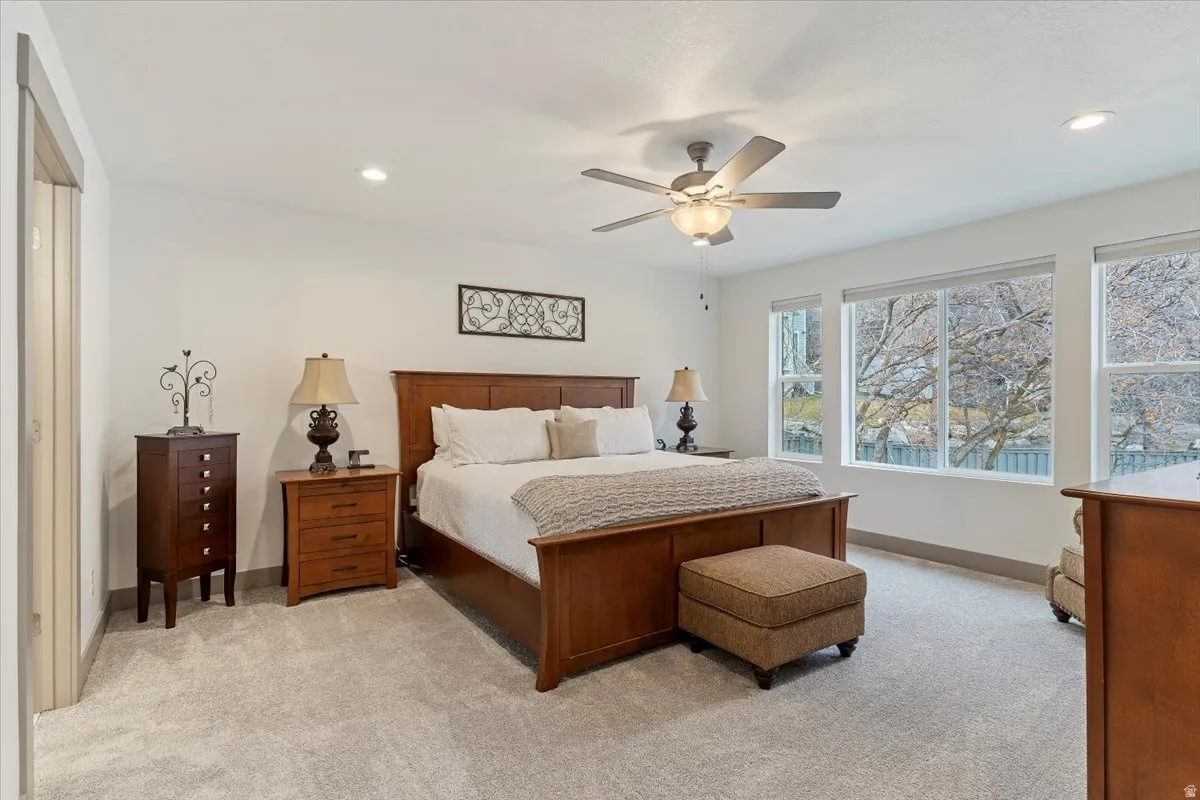 Spacious bedroom featuring light-toned carpeting, recessed lighting, a ceiling fan with integrated lighting, and multiple large windows providing abundant natural light