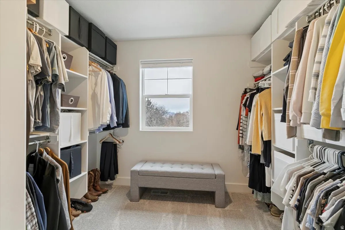 Spacious walk-in closet featuring white built-in shelving, multiple hanging rods, a central window, and light gray carpet flooring
