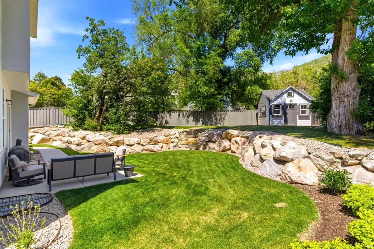 Expansive backyard featuring a manicured lawn, a rock retaining wall, a dedicated patio area, mature trees, and a detached shed