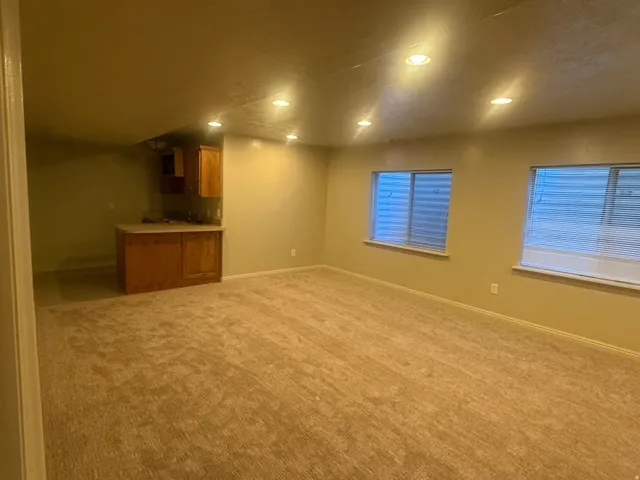 Carpeted flooring throughout, recessed ceiling lighting, two windows with blinds, and a built-in wet bar with wood-finish cabinetry