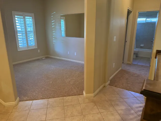 Carpeted room with a window featuring white plantation shutters, tile flooring, and a full bathroom with a tiled shower and white bathtub