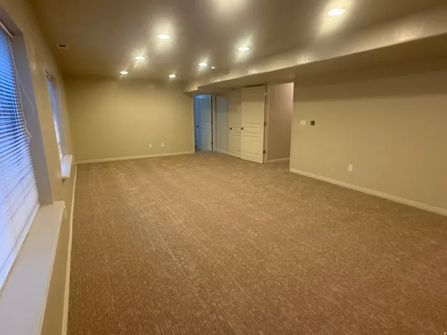 Expansive carpeted room with multiple recessed lighting fixtures, light beige walls, and two windows with horizontal blinds