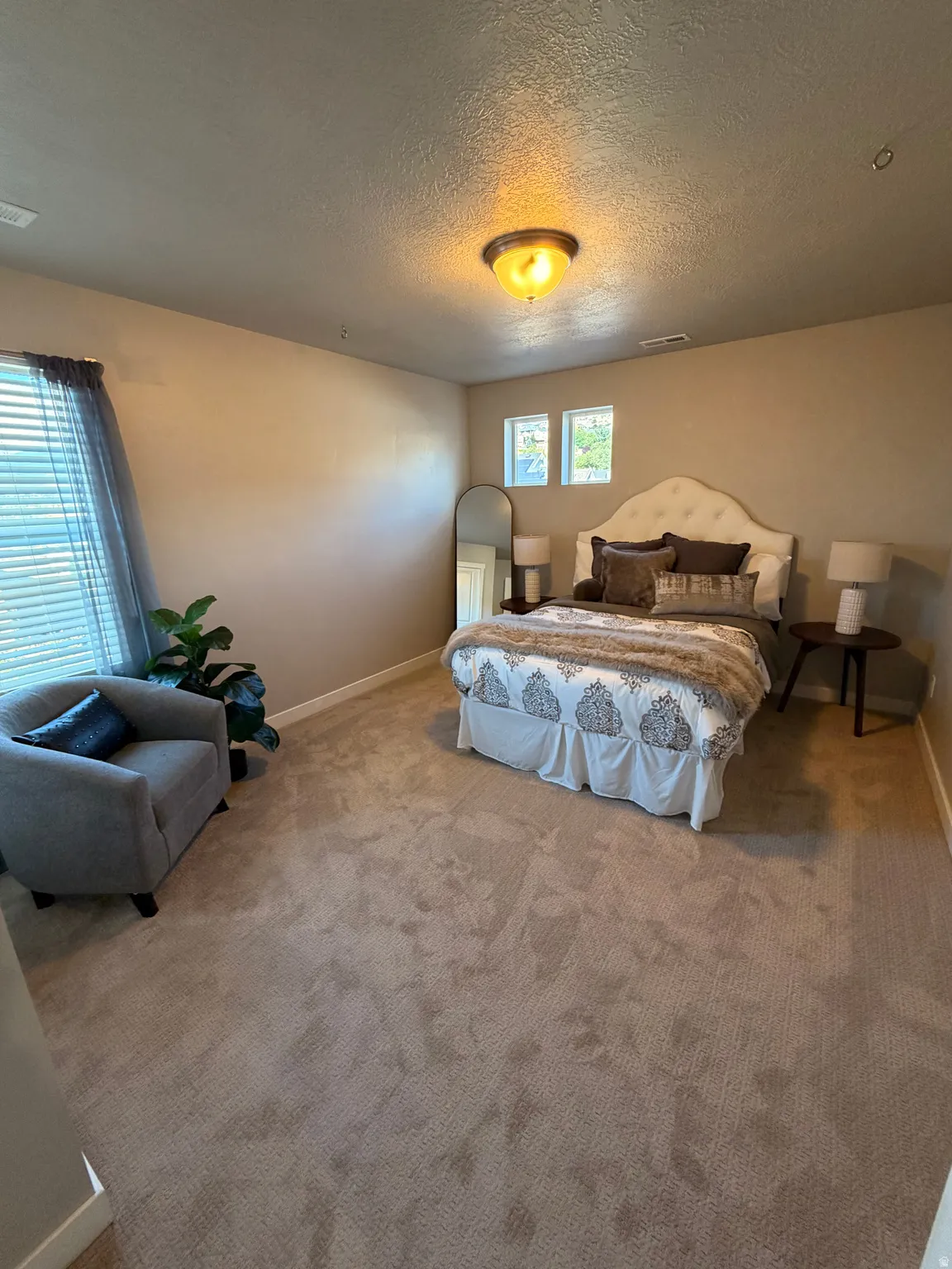 Carpeted room featuring a ceiling-mounted light fixture, two windows with blinds, a built-in upholstered headboard, and a neutral wall color