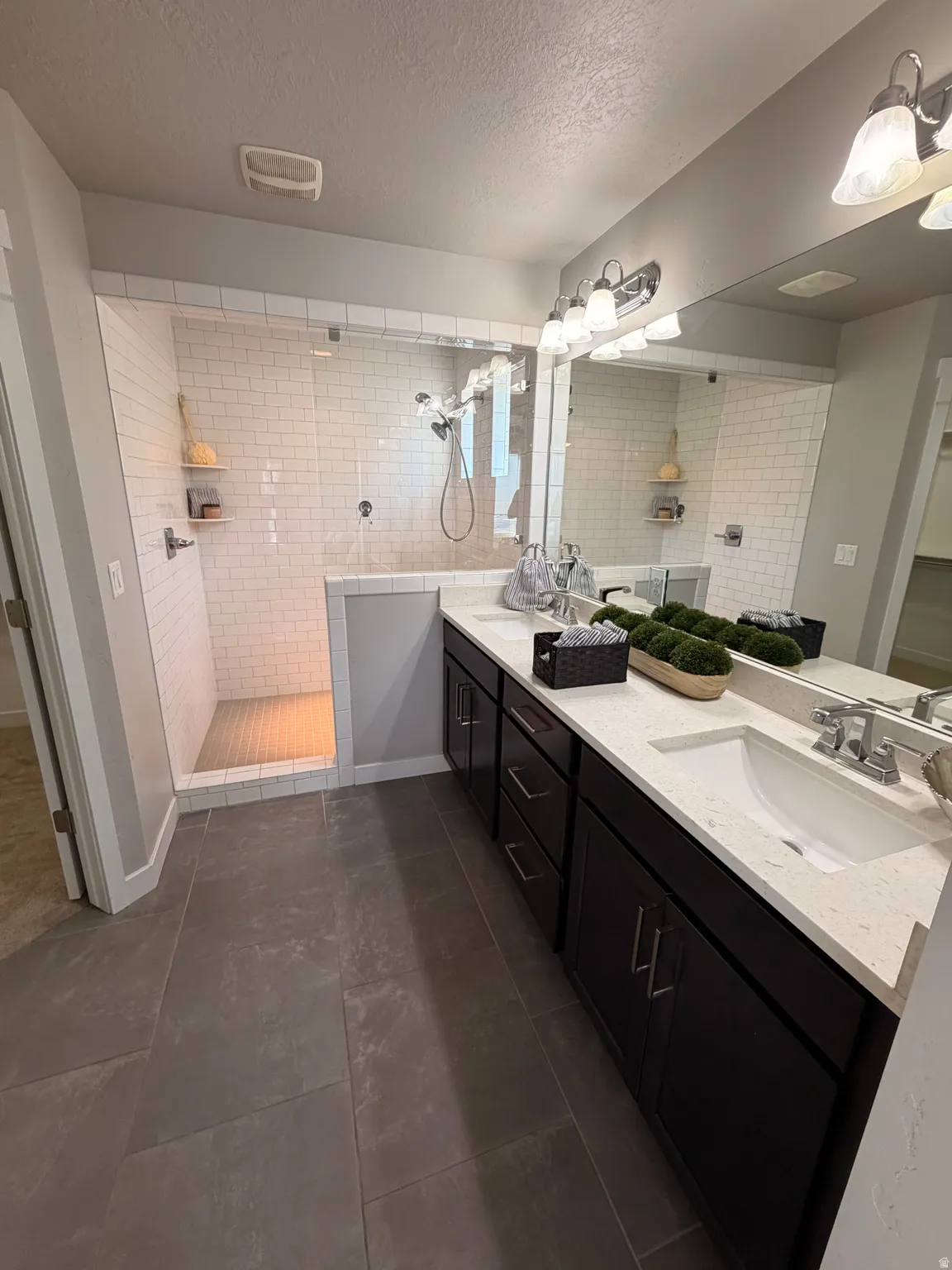 Spacious bathroom featuring a double vanity with dark cabinetry and light-toned countertops, a large mirror, and a walk-in shower with subway tile surround and glass enclosure