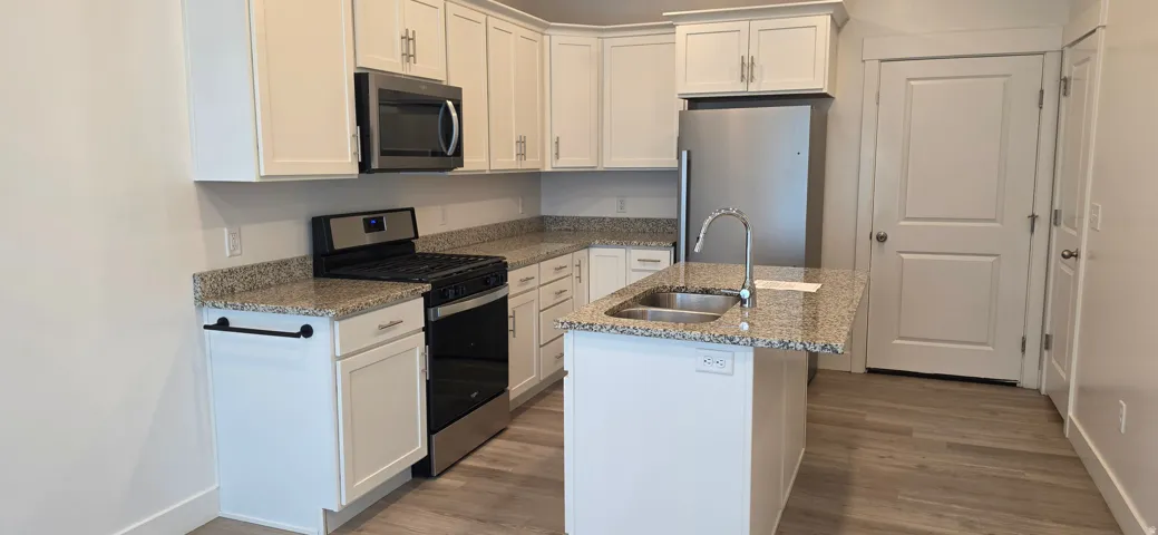 Contemporary kitchen featuring white cabinetry, granite countertops, stainless steel appliances, a center island with a double basin sink, and wood-finish flooring