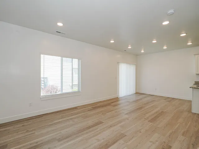 Spacious room featuring wood-finish flooring, white walls, recessed ceiling lighting, a large window with blinds, and a sliding glass door