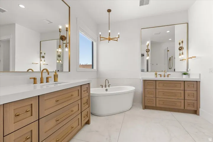 Freestanding soaking tub with brushed gold fixtures, dual wood-finish vanities with white countertops, and large framed mirrors