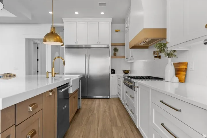 Gourmet kitchen featuring a large island with a farmhouse sink, stainless steel appliances, wood-finish flooring, white shaker cabinetry, and a brass range hood